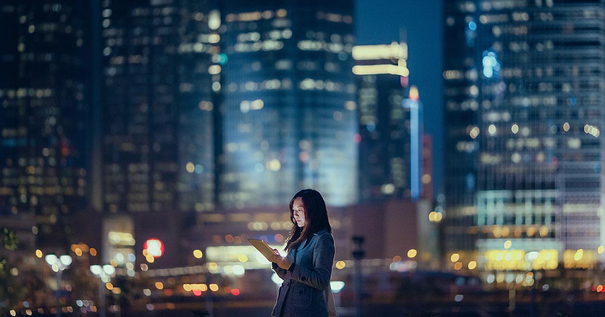 Woman in office at night using digital tablet, standing against illuminated highrise corporate buildings, device, individual, city